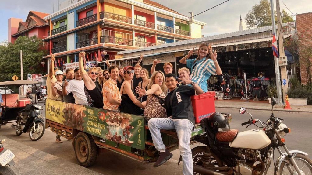 A group of international backpackers riding a tuk-tuk together in Bangkok, Thailand, smiling and waving as they explore local markets and experience social, community-driven travel.