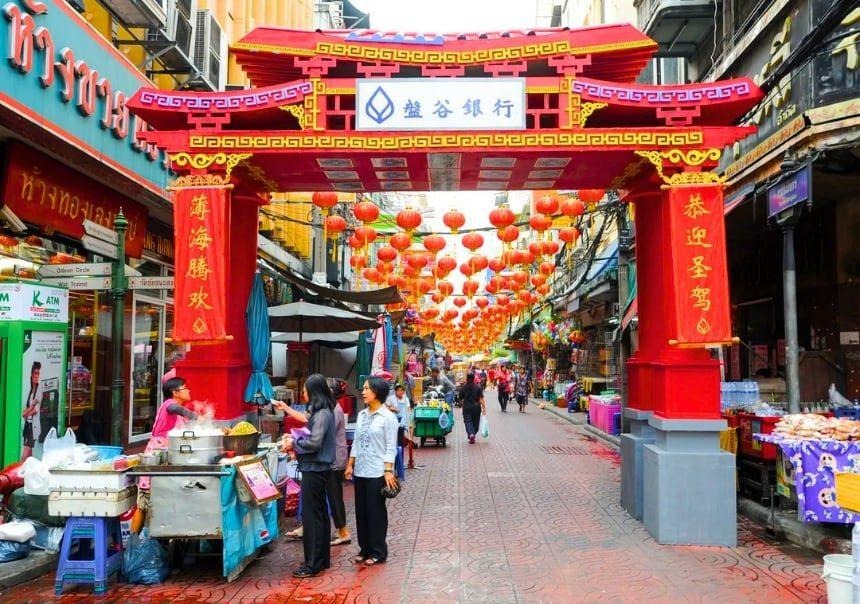 Red entrance arch, lanterns, vendors, and shoppers along Sampheng Market in Bangkok Chinatown